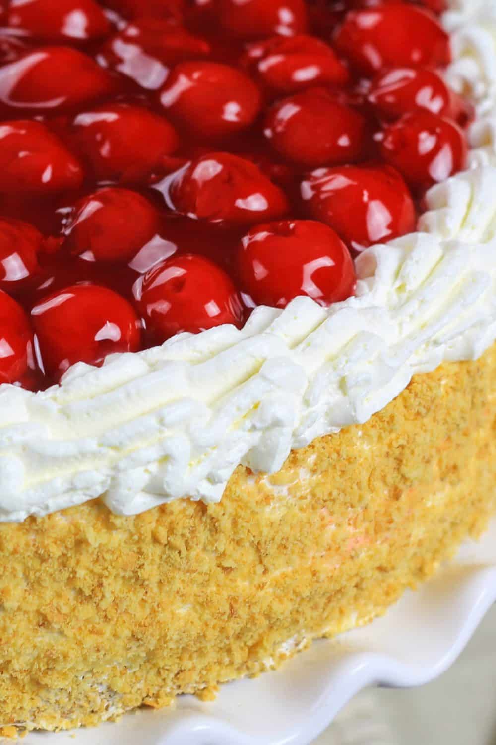 This Cherry Jello Cake Recipe is one of my favorite summertime desserts! A homemade cake and layers of sweet cherry filling make it a hit you feel good serving. #jellocake #cherryjello #spongecake #cherrypiecake #cherrycake Up close picture of a completed cherry jello cake recipe with the cake sitting on a white cake plate and topped with cherry pie filling, cream cheese filling, and a coating of cake crumbs around the edges