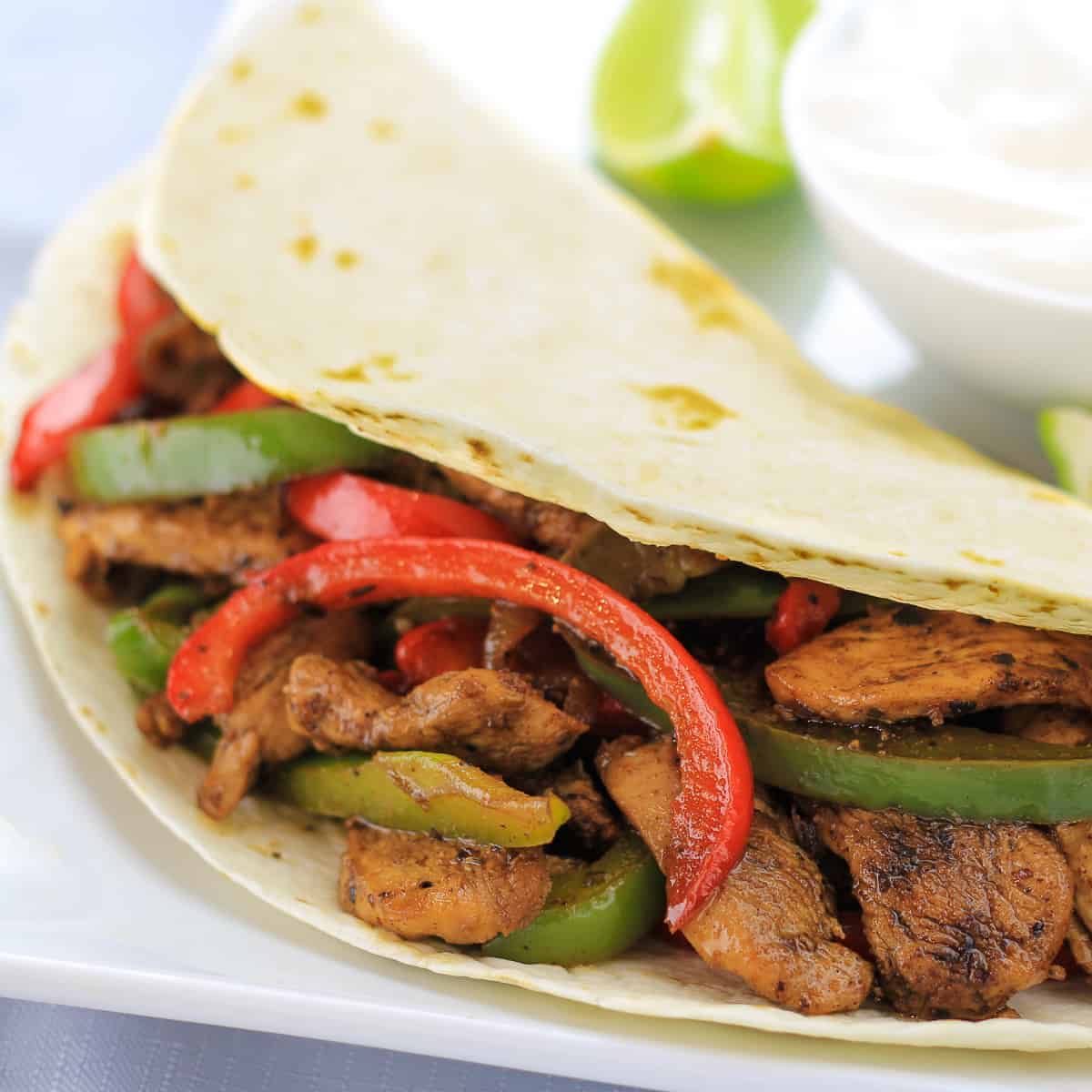 chicken fajitas on a white plate with a bowl of sour cream and to limes next to it