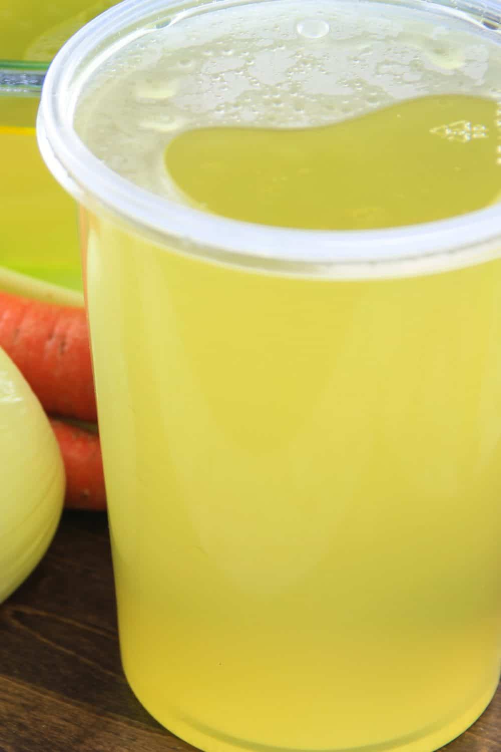 Homemade chicken broth is so simple to make and the flavors cant compare to the storebought kind. Make a big batch and use it for delicious soups, stews, all kinds of sauces. #chickenbroth #homemadebroth #chickenstock #bonebroth a close up image of homemade chicken broth in a large plastic tub with onion and carrots in the background