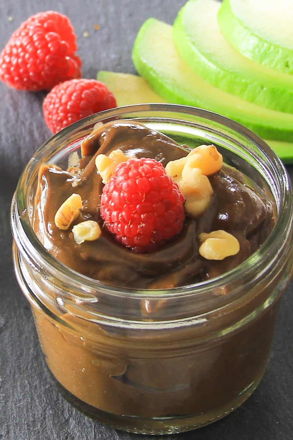 This Paleo Chocolate Pudding is shockingly easy to make and packs smooth creamy chocolate flavor you are looking for while staying Paleo friendly. #paleochocolate #chocolatepudding #healthypudding #paleodessert Small mason jar full of Paleo chocolate pudding topped with chopped walnuts and a raspberry sitting on a dark wooden surface with raspberries and avocado in the background