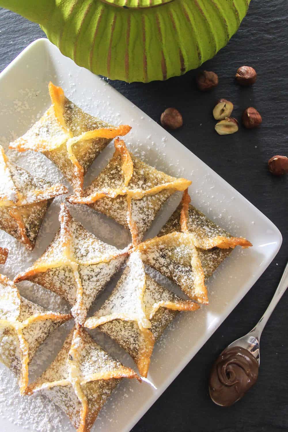 Nutella filled wonton recipe on white platter topped with powdered sugar and displayed on a dark surface with green teapot in background