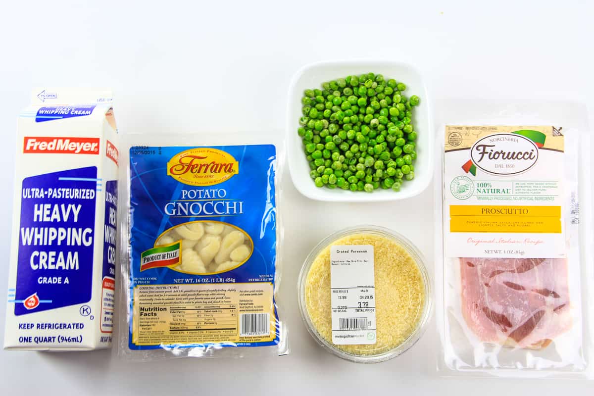 Ingredients for potato gnocchi recipe laying on a flat white surface including heavy cream, gnocchi, Parmesan cheese, green peas, and prosciutto
