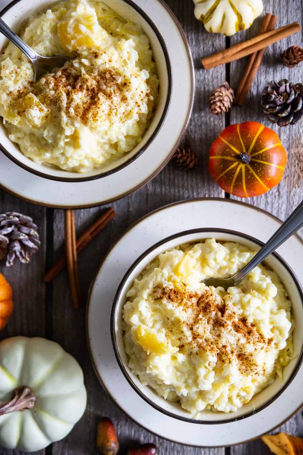 This simple rice pudding recipe is the perfect comfort dish for fall since its loaded with cubes of sweet spaghetti squash. The flavors are truly amazing! #ricepudding #fallrecipe #kasha #Ukrainianporridge an overhead image of two bowls of rice pudding with cinnamon sticks and mini pumpkin beside them on a dark wooden background