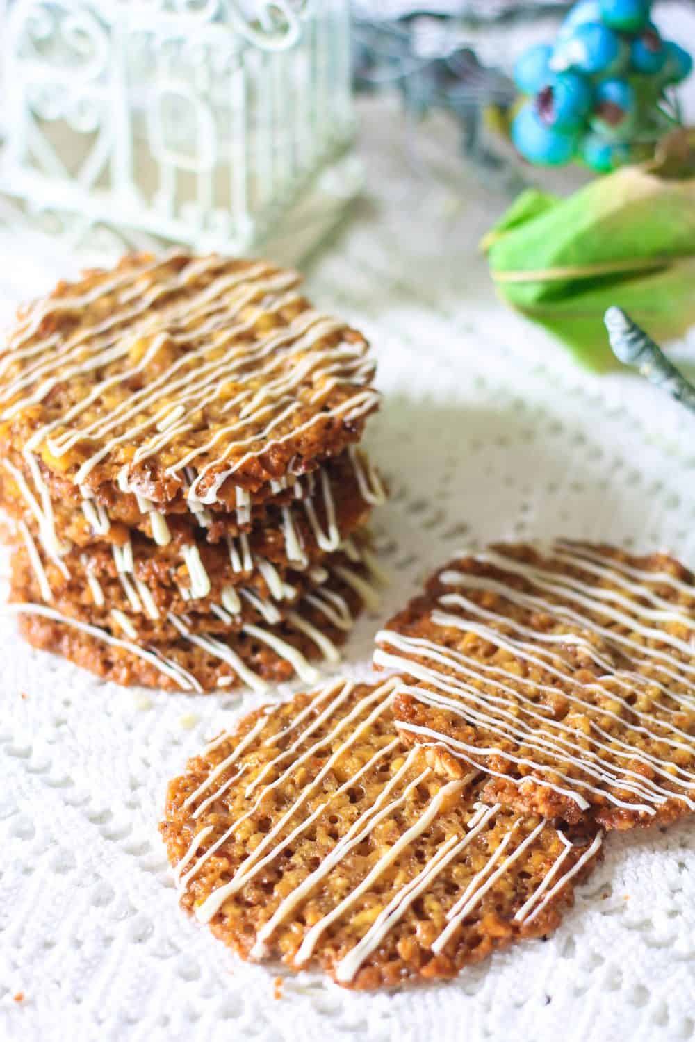 Florentine Cookies are the perfect delicate balance of crunchy sweetness that melts in your mouth. This simple recipe will become a favorite. #Florentinecookies #lacecookies #almondcookies #thincooking #Florentines A stack of florentine cookies with white chocolate drizzles on top of each cookie laying on a white lace tablecloth with a stack of cookies as well as a few laying flat