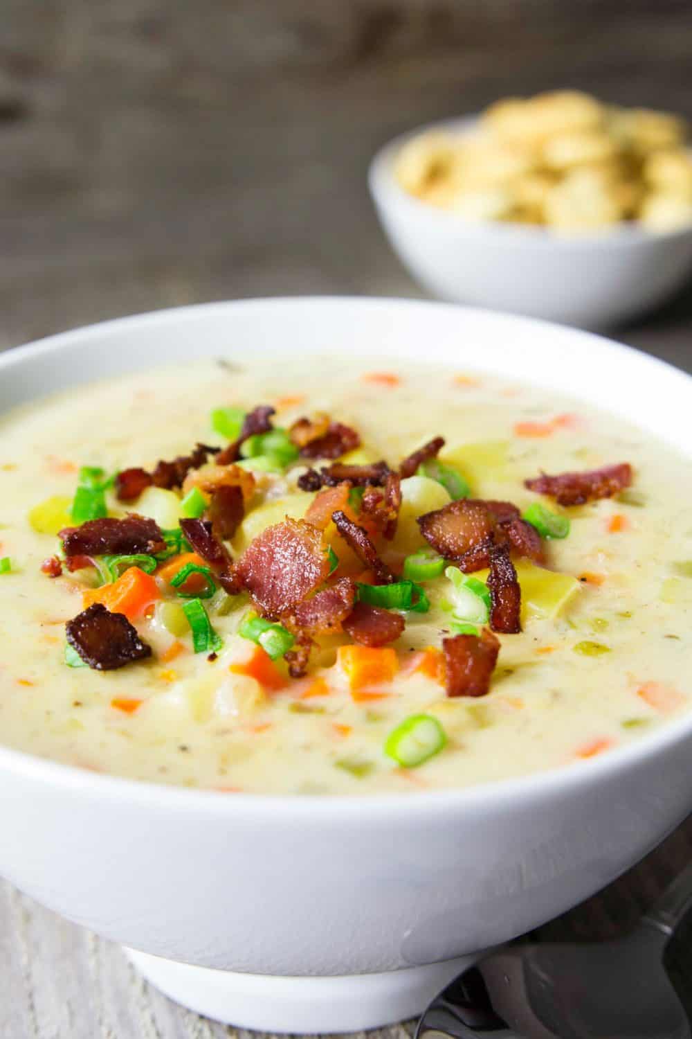 This Clam Chowder Soup recipe is a perfect meal that brings together a potato and cream base and the saltiness of clams with a light smoky bacon flavor. #clamchowder #chowder #seafoodchowder #clamsoup #seafoodsoup #creamysoup A large white bowl on a wooden table full of clam chowder soup that has been topped with bacon crumbles and diced green onions