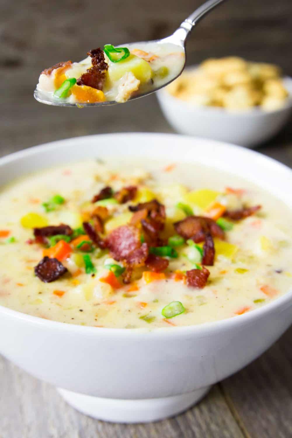 This Clam Chowder Soup recipe is a perfect meal that brings together a potato and cream base and the saltiness of clams with a light smoky bacon flavor. #clamchowder #chowder #seafoodchowder #clamsoup #seafoodsoup #creamysoup Large white bowl of clam chowder sitting on a wooden surface with a spoon hovering over the top full of soup