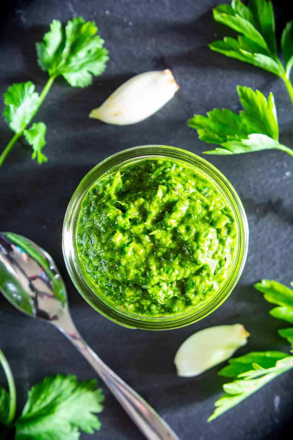 Chimichurri sauce in bowl with garlic cloves, parsley, and a spoon in the background.