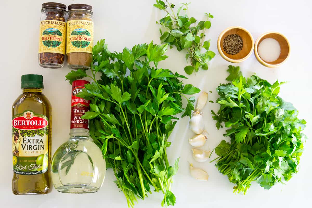 Ingredients for chimichurri sauce, including oil, vinegar, parsley, oregano, cilantro, cumin, red pepper flakes, garlic, and salt & pepper.