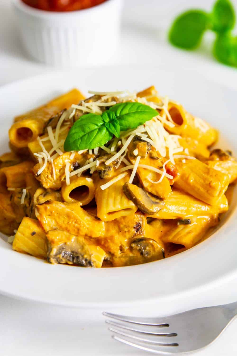 Up close image of creamy chicken mushroom pasta recipe in white bowl on table with grated parmesan cheese and basil as a topping.