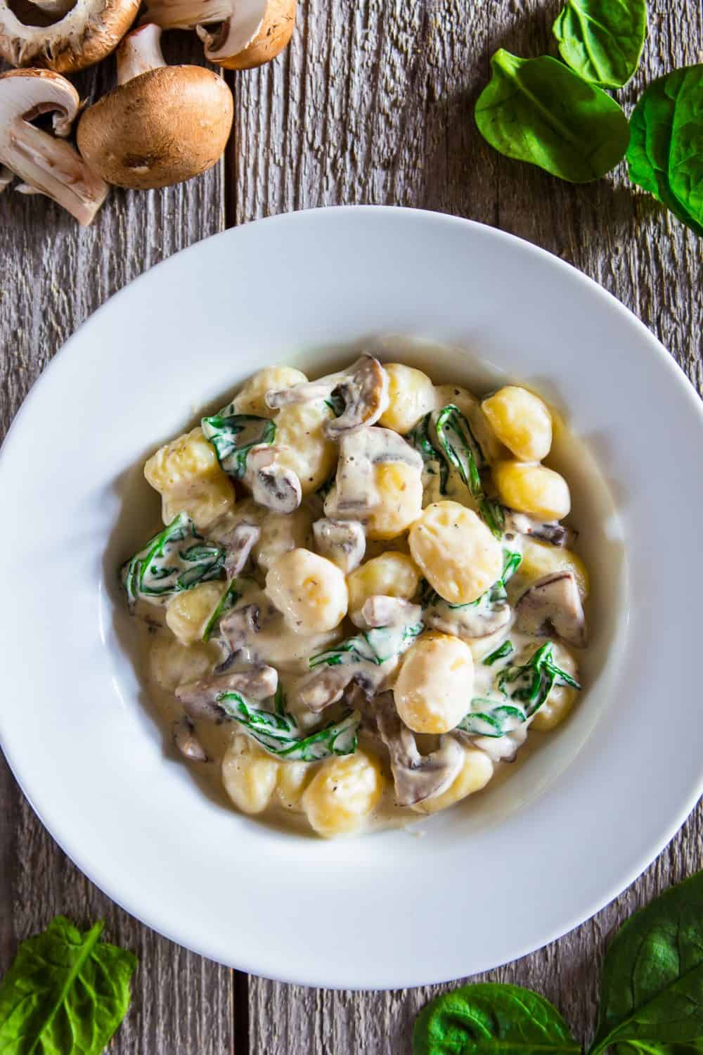 creamy mushroom gnocchi with spinach in a white bowl with fresh spinach and raw mushrooms beside it
