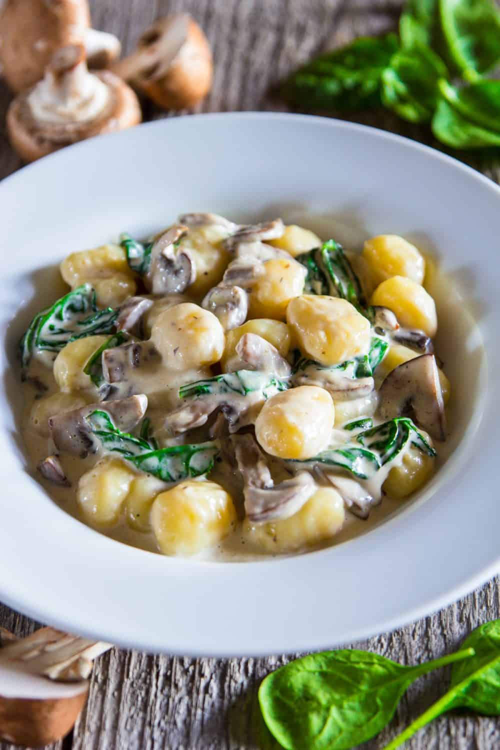 creamy mushroom gnocchi with spinach in a white bowl with fresh spinach and raw mushrooms beside it