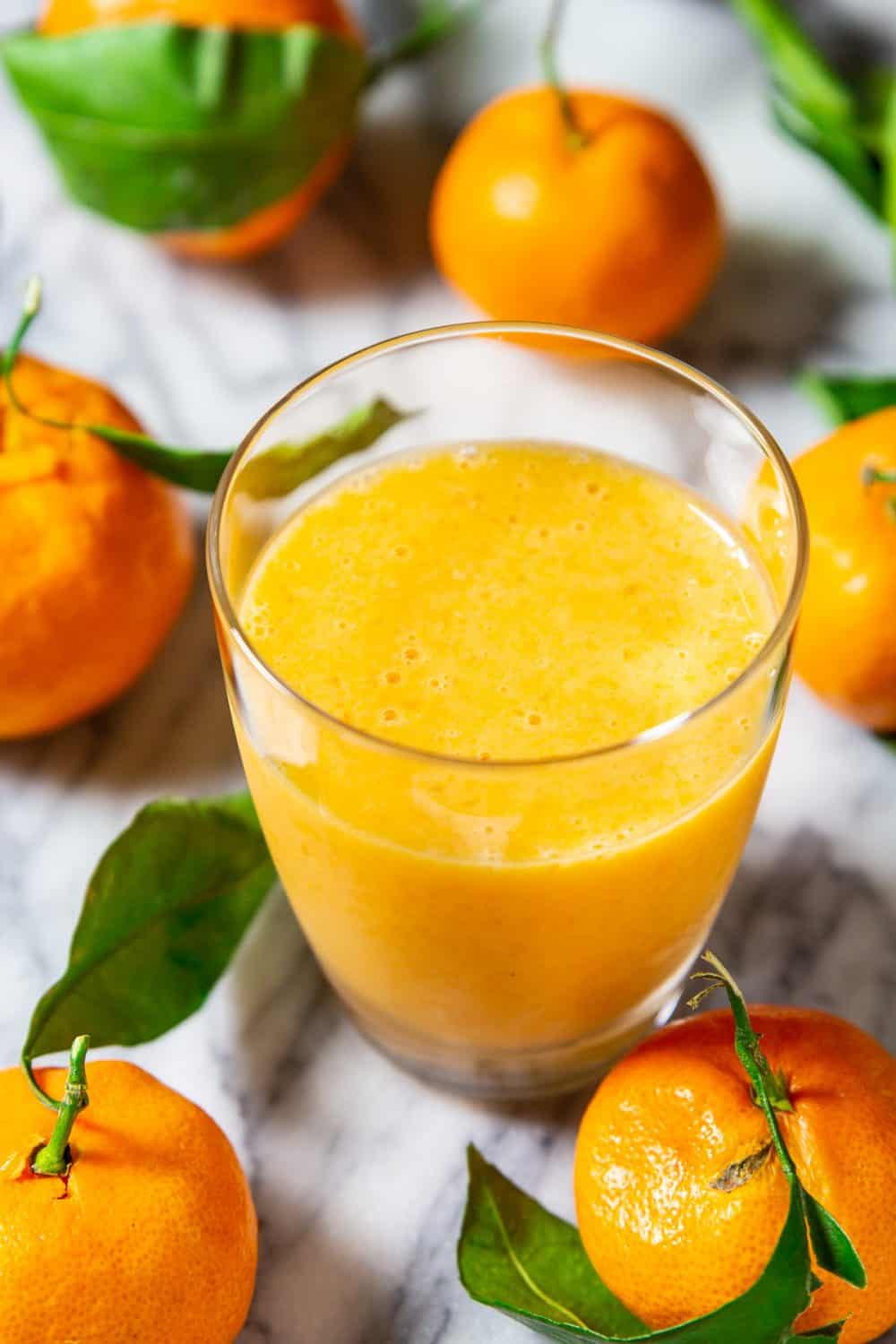 Image of mandarin banana smoothie in a small glass without a straw sitting on a marble counter surrounded by mandarins
