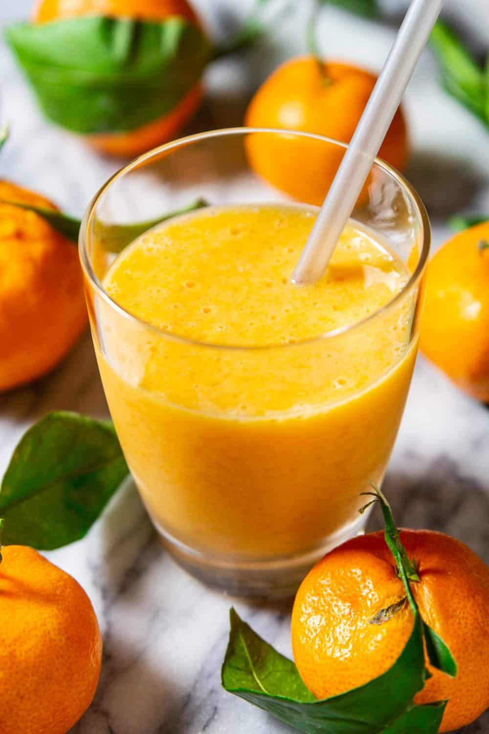 Up close image of mandarin and banana smoothie recipe in a small glass with straw on a marble counter surrounded by fresh manadarins