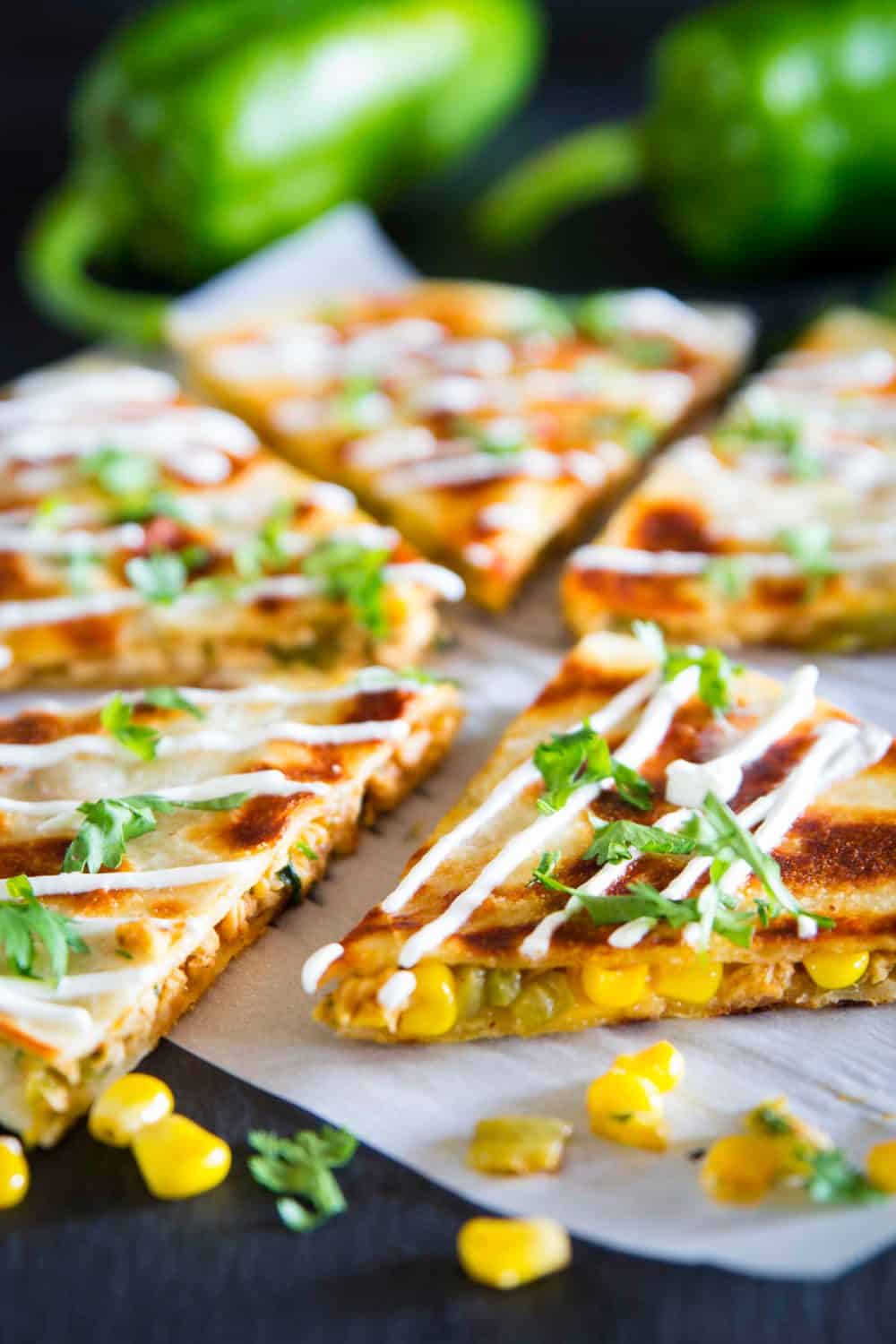 These easy chicken quesadillas are loaded with Mexican cheese, hatch chilies, and juicy rotisserie chicken. Its the perfect recipe for busy weeknights! #chickenquesadillas quesadilla #mexicanrecipe #mexicanfood #30minutemeal a close up shot of sliced chicken quesadilla with a corn, sour cream, and chopped cilantro on top