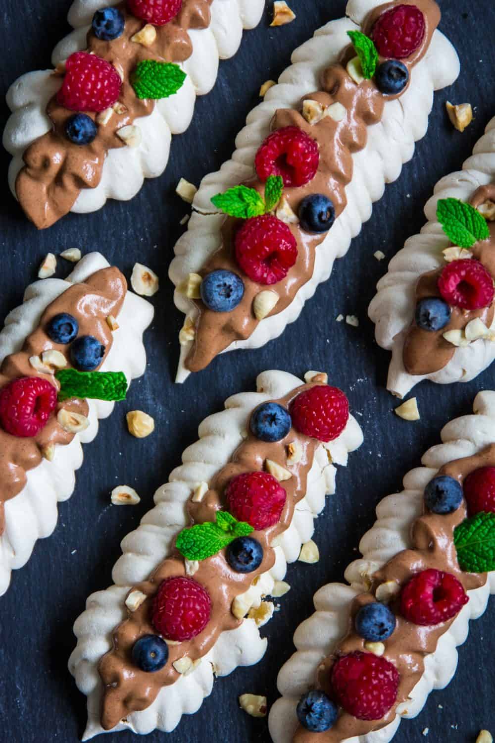 These meringue bites are topped with delicious chocolate cream, toasted hazelnuts, and fresh berries. It's an elegant dessert that everyone loves! #meringuecookies #meringuebites #russiandessert #meringuedessert overhead image of image of homemade meringue cookies with berries, hazelnuts and chocolate cream on top