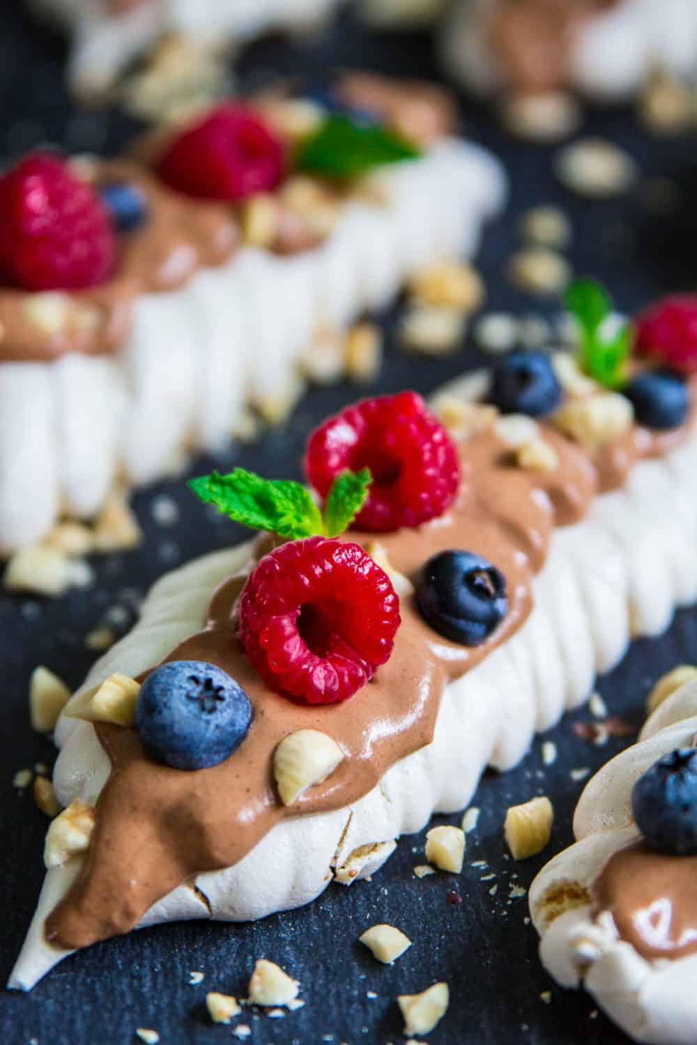 These meringue cookies are topped with rich chocolate cream, chopped hazelnuts, and fresh berries. It's a delicate dessert that everyone loves! #meringuecookies #meringuebites #russiandessert #meringuedessert close up image of homemade meringue cookies with berries and chocolate cream on top