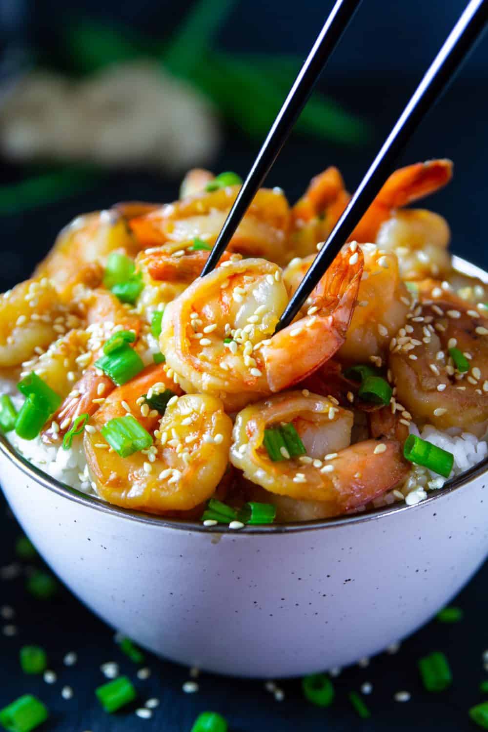 garlic ginger shrimp with green onion and sesame seeds on top in a white bowl with black chop sticks picking up the shrimp