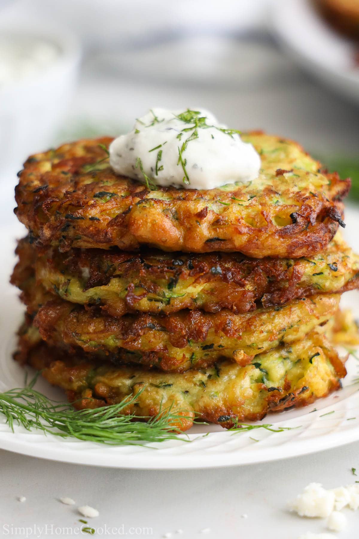 This zucchini fritters recipe is not as basic as you think. The addition of crumbled feta cheese gives them a cheesy Greek twist that is oh so irresistible. #zucchinifritters #zucchini #appetizers #summerfood #zucchinipatties #fritters Stack of Zucchini Fritters with a dollop of tzatziki sauce on top and some fresh dill