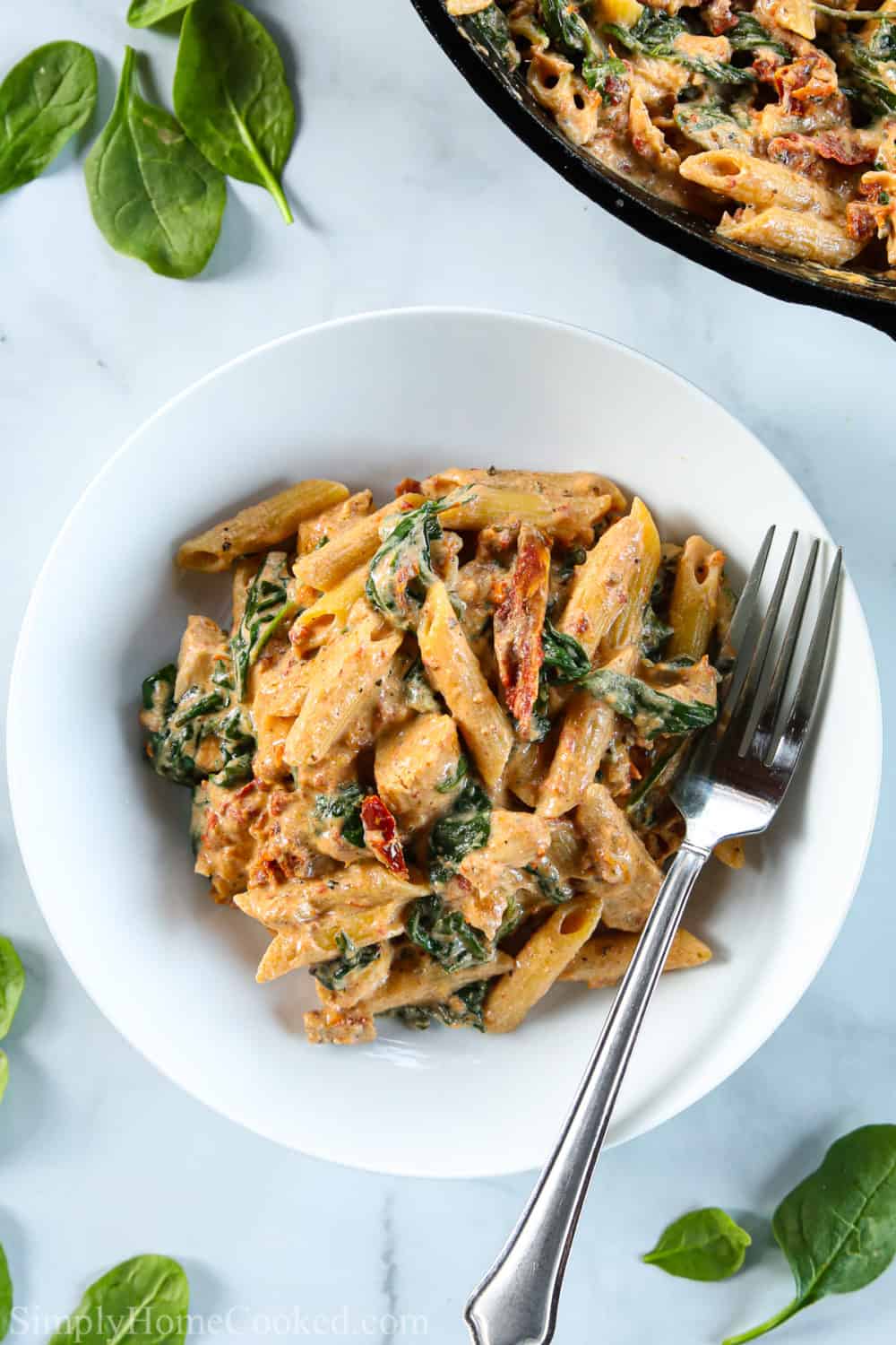 Plate of Creamy Tuscan Chicken Pasta with a fork, and spinach and a skillet of pasta in the background.