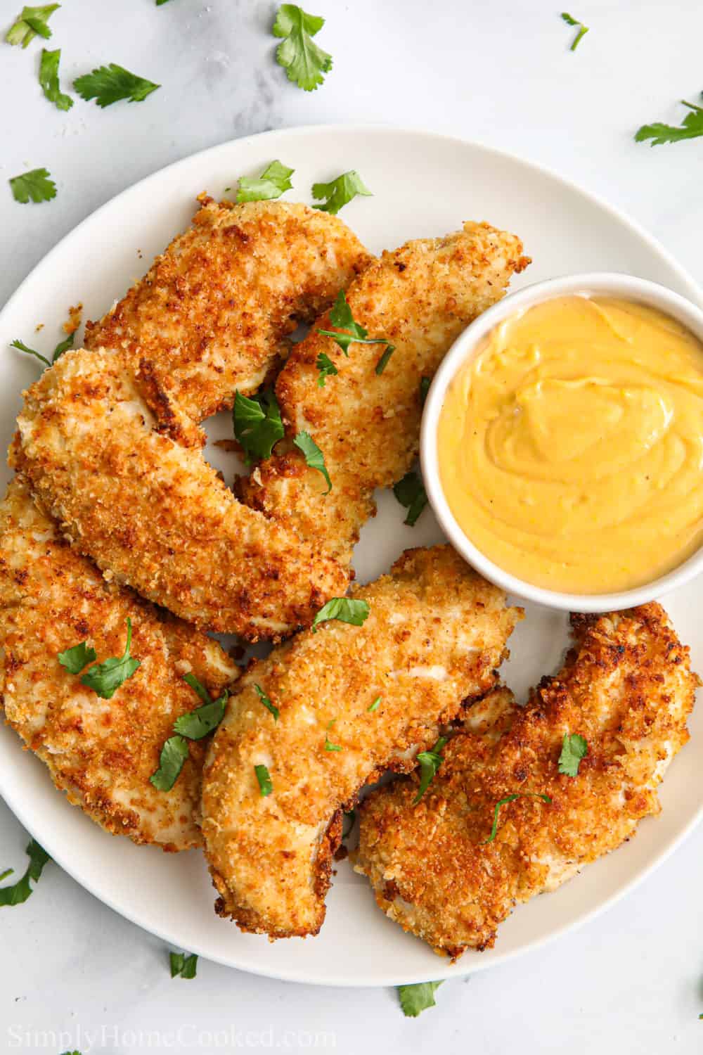 White plate with Breaded Air Fryer Chicken Tenders, garnished with parsley, and with a side of dipping sauce.