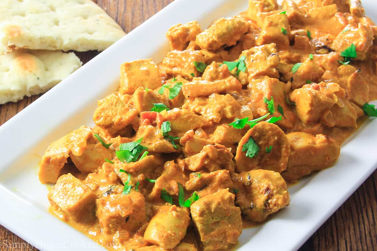 This Indian Butter Chicken is so delicious and easy to make. It's full of flavor and makes a filling and yummy dinner. #chickenbuttersauce #marinatedbutterchicken #bestindianbutterchickenrecipe #easyhomemadebutterchicken White plate filled with Indian Butter chicken with some parsley on top and bread to the side.