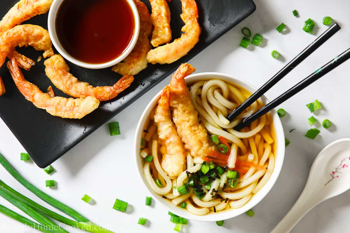 Bowl of udon soup with chopsticks and shrimp tempura in it, and a plate of shrimp tempura and dipping sauce to the side; green onions and a spoon next to them.