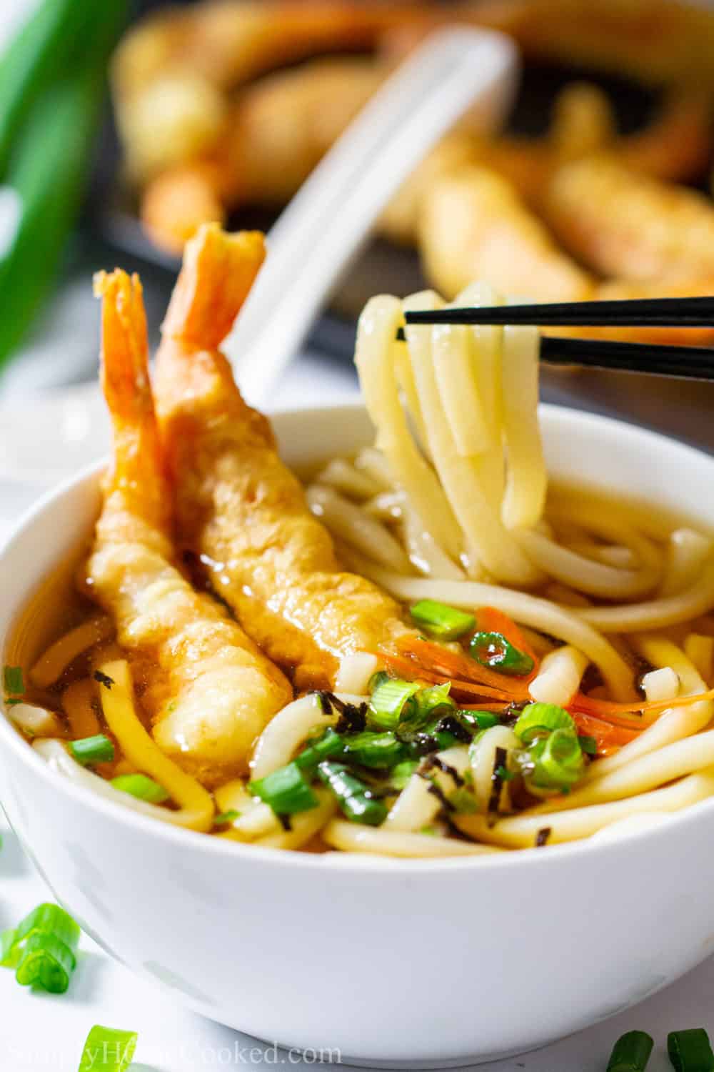 Close up of a bowl of udon soup with shrimp tempura on top , chopsticks lifting the noodles, and shrimp tempura in the background.