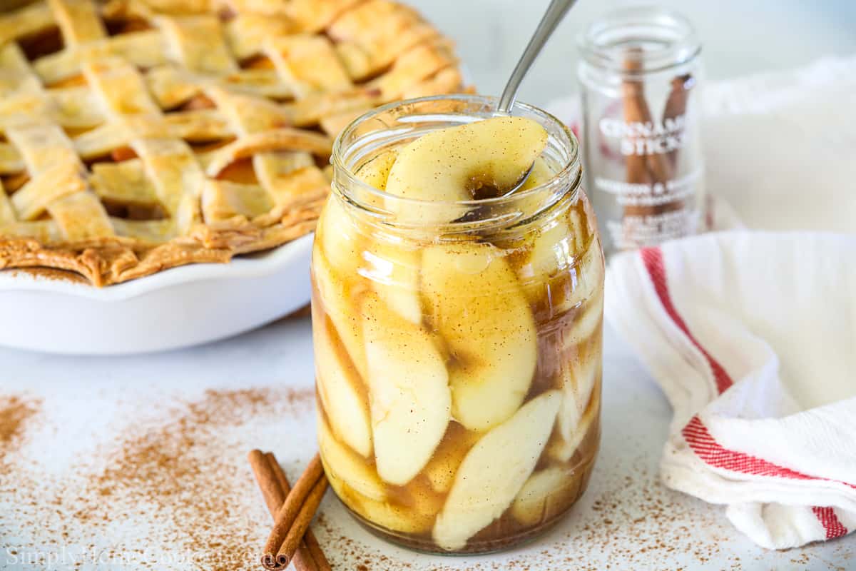 This Homemade Apple Pie is sweet and cinnamony and so delicious. Nothing tastes better than apple pie right out of the oven! #applepiefromscratch #scratchapplepie #scratchdesserts #homemadedessert Apple pie filling in a jar with a spoon, and an apple pie in the background with cinnamon sticks and a towel.
