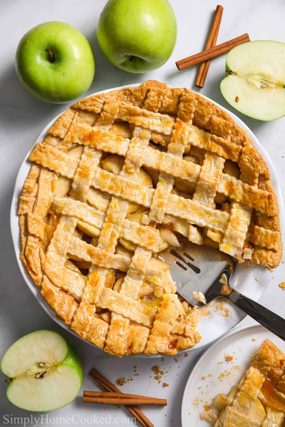This Homemade Apple Pie is made with sweet cinnamon apples and a beautiful lattice pie crust. Golden brown, flaky, and delicious, it is perfect for company. #homemadepie #homemadeapplepie #applepie #applepierecipe Homemade apple pie with a slice cut out and some green apples and cinnamon sticks nearby.