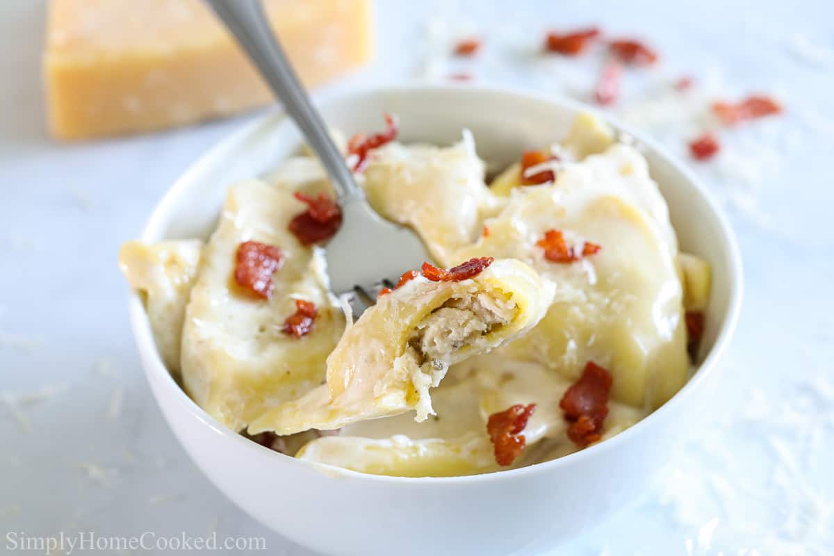 Chicken and Cheese Ravioli with a cream sauce and bacon bits in a white bowl with a fork in it, cheese and bacon in the background.