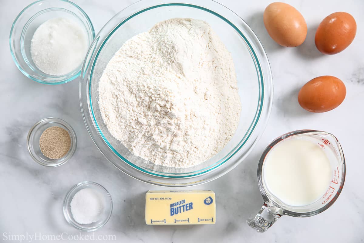 Ingredients for Soft Brioche Dinner Rolls, including flour, sugar, yeast, brown eggs, butter, milk, and salt on a white background.