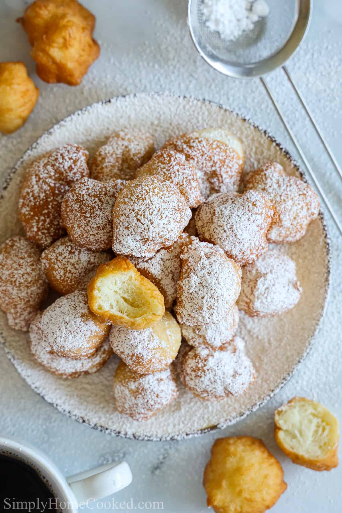 Plate of powdered sugar dusted zeppoles with one bitten, and some zeppoles and a small sieve to the side on a white background.