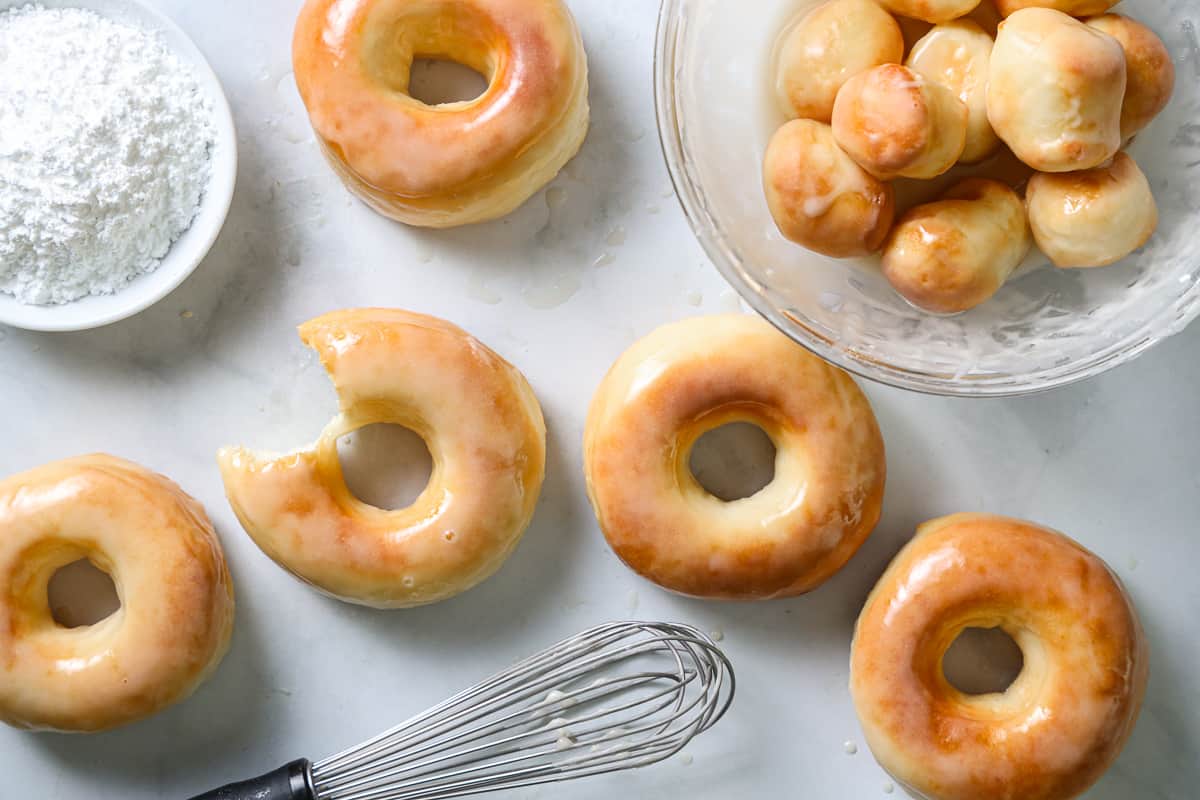 Glazed Air Fryer Donuts on a white background with a bowl of donut holes and a bowl of powdered sugar and a whisk nearby.
