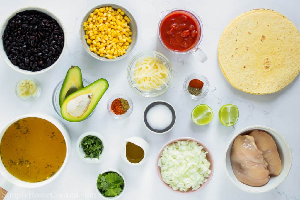 Ingredients for Chicken Tortilla Soup, including corn tortillas, chicken tenders, black beans, frozen corn, diced tomatoes, chicken broth, garlic, onion, shredded cheese, lime, salt, olive oil, cilantro, oregano, cumin, chili powder, and smoked paprika, ona  white background.