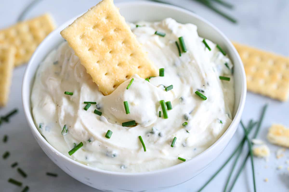 Close up of Garlic Cream Cheese Dip with a cracker in it and chopped chives on top, more crackers and chives in the background.