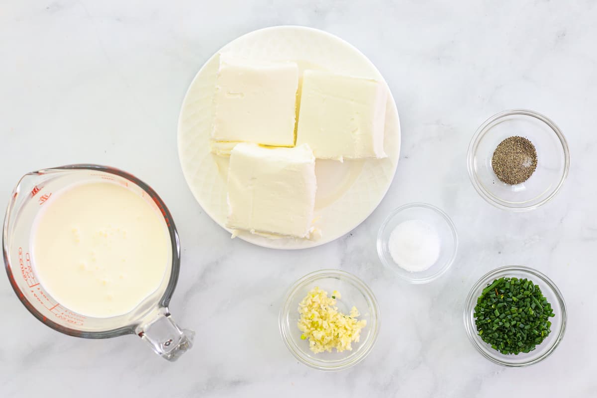 Ingredients for Garlic Cream Cheese Dip, including cream cheese, heavy cream, chives, salt, pepper, and minced garlic, on a white background.