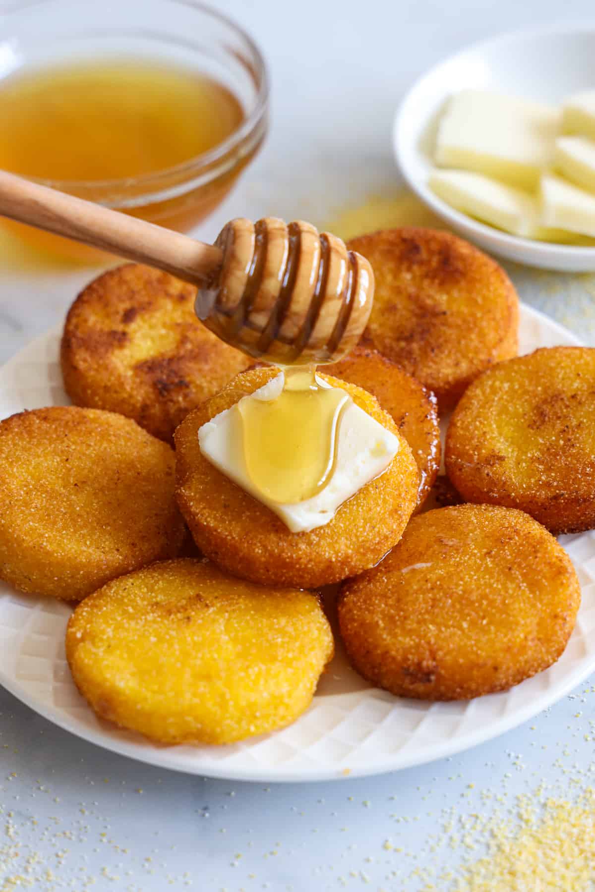 This Southern Hot Water Cornbread is dense, moist, & delicious. Fried to a golden brown, it is allergy-friendly, flavorful, & beyond tasty! #hotwatercornbread #cornbread #southerncornbread #southernhotwatercornbread Southern Hot Water Cornbread on a plate with a pat of butter and honey being dribbled on it, honey and butter in the background.
