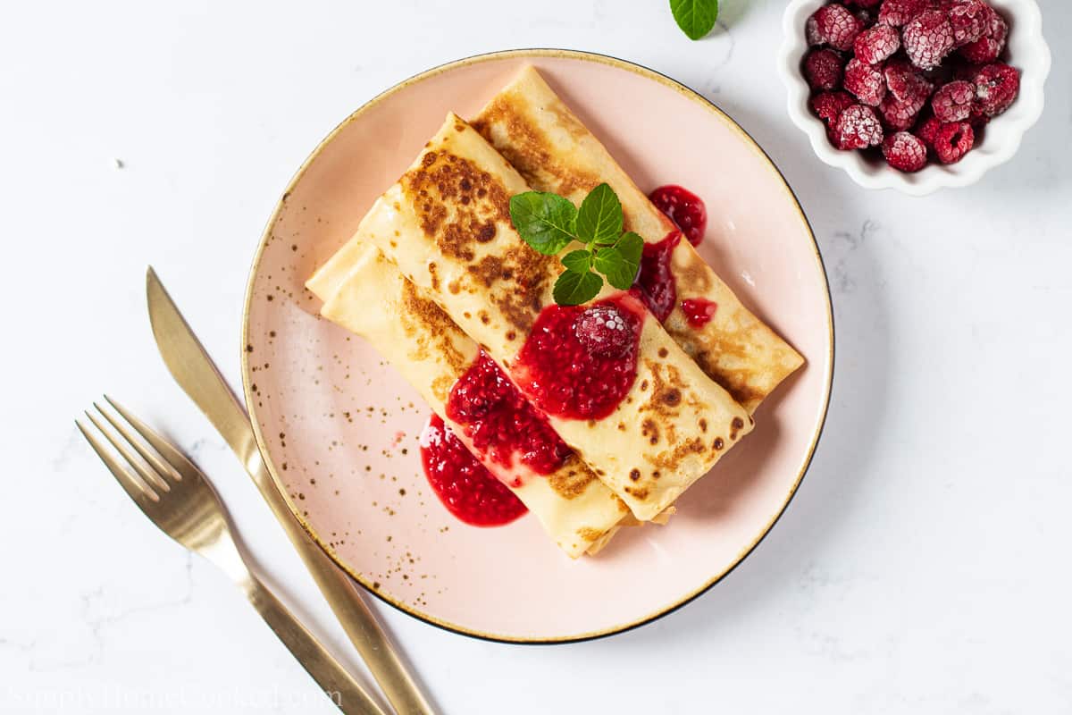 Overview Cheese Blintz on a plate with berry sauce on top with garnish and a fork and knife nearby, and a bowl of raspberries to the side.