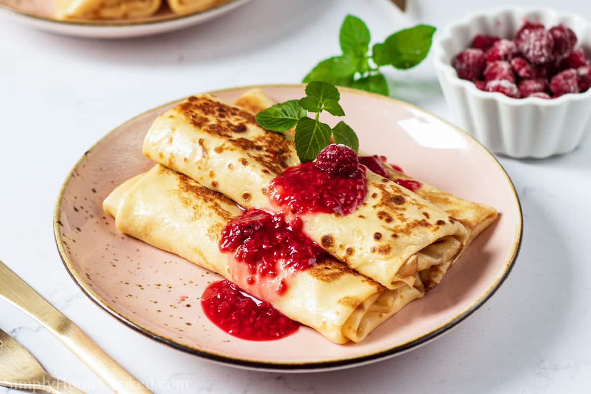 Cheese Blintz on a plate with berry sauce and garnished on top, with silverware nearby and a bowl of raspberries in the background.