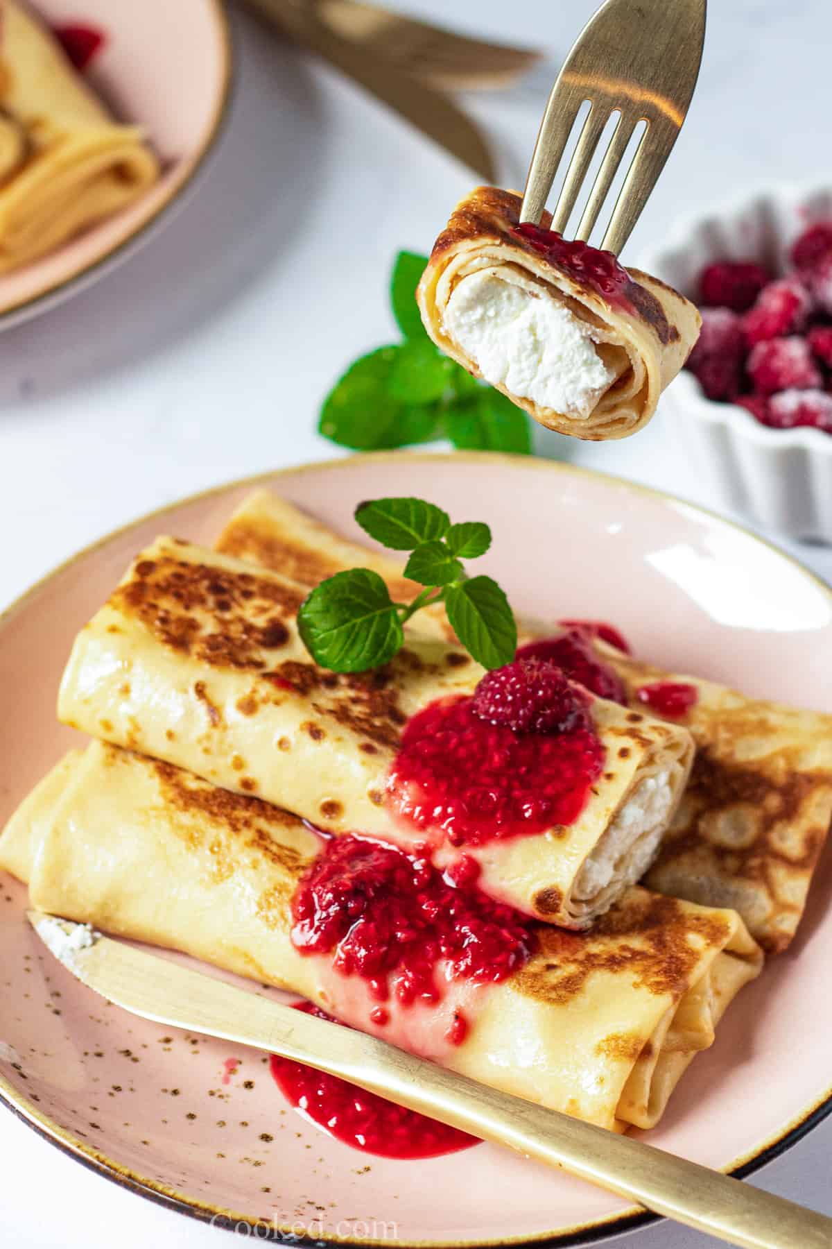 Closeup of a Cheese Blintz with berry sauce and a garnish on top, a knife next to them on the plate and a fork with a piece of blintz on it.