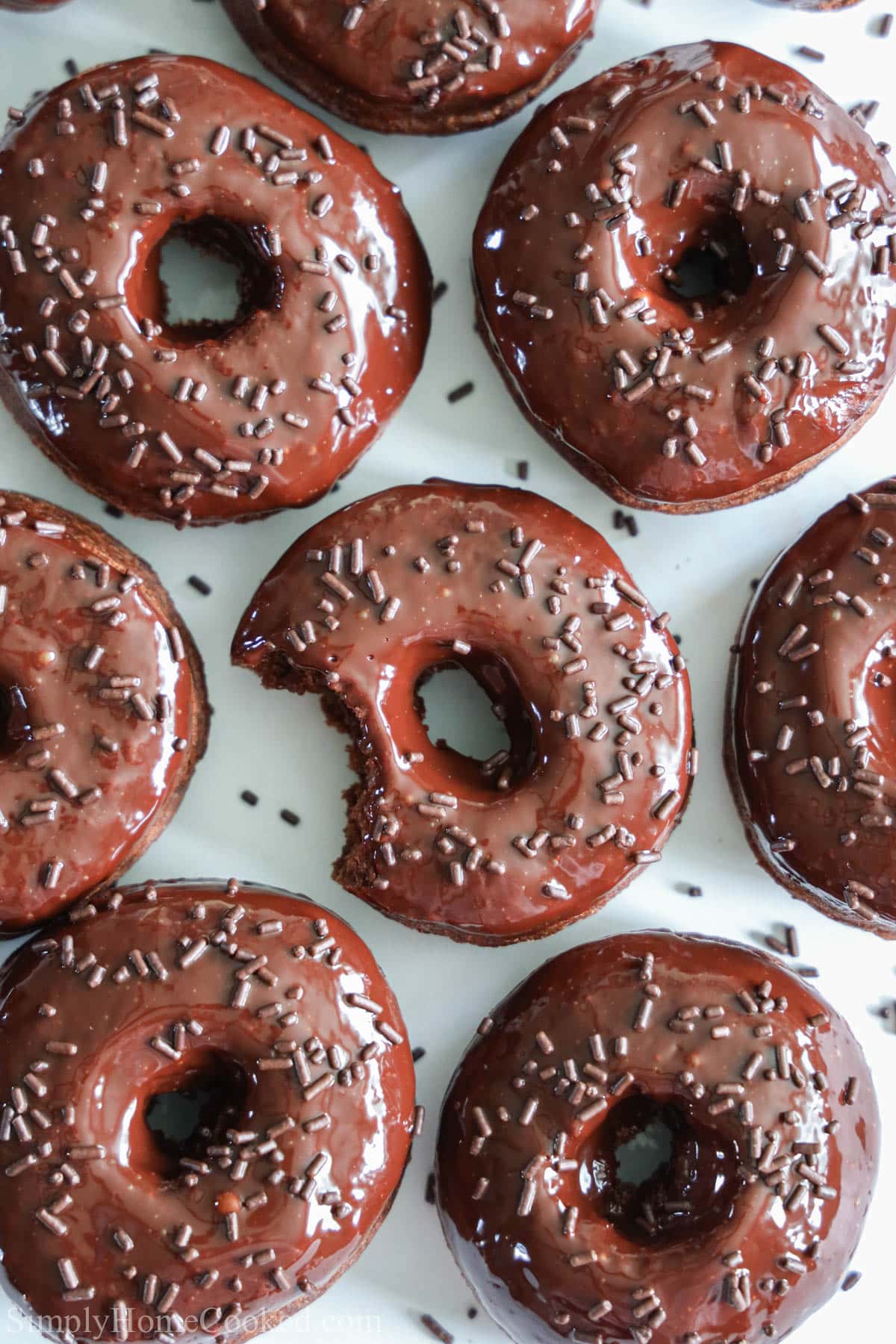 These Chocolate Donuts are so delicious and moist. Sweet and tangy with a smooth glazed top, this recipe is too yummy not to try! #bestdonuts #bestchocolatedonuts #bestchocolateglazeddonuts #bestglazedchocolatedonuts Chocolate Donuts covered in sprinkles, one missing a bite