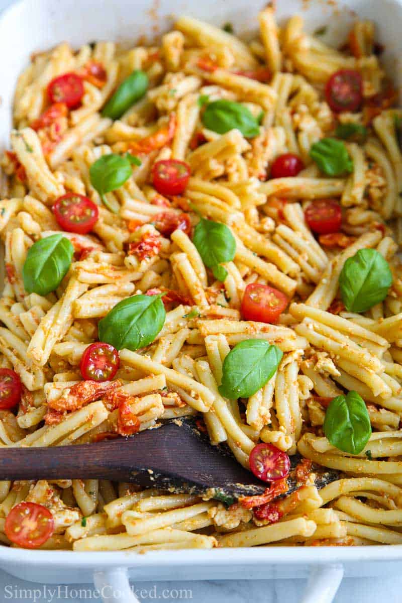 Close up of a baking pan of Baked Feta Pasta being mixed with a wooden spoon and garnished with basil leaves.