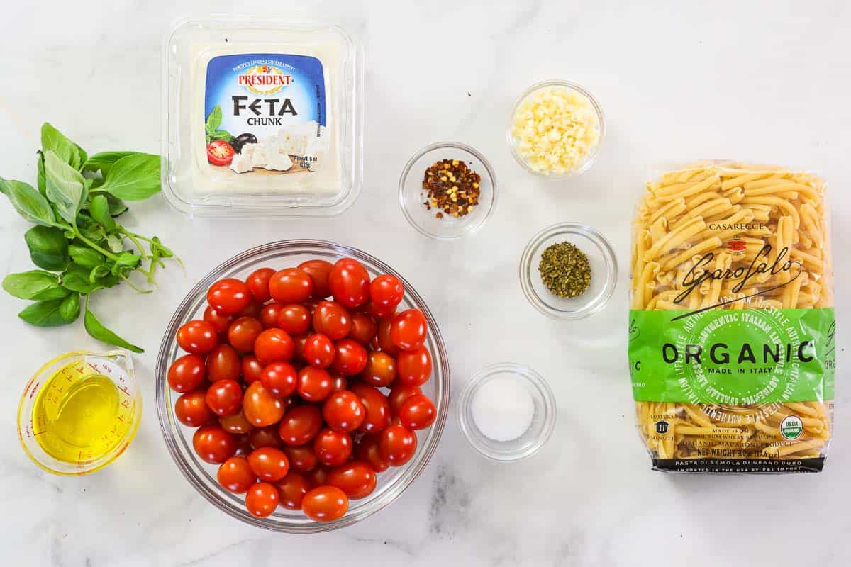 Ingredients for Baked Feta Pasta, including dry pasta, feta cheese, cherry tomatoes, basil leaves, olive oil, salt, pepper, garlic, and red pepper flakes.