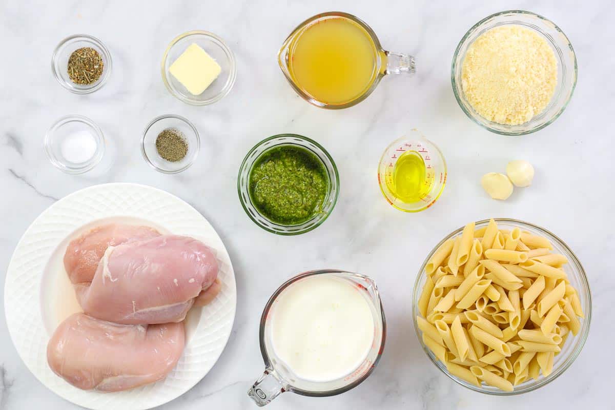 Ingredients for Creamy Chicken Pesto Pasta, including chicken breasts, penne pasta, Parmesan cheese, basil pesto, chicken broth, oil, garlic cloves, salt, pepper, butter, and heavy cream.