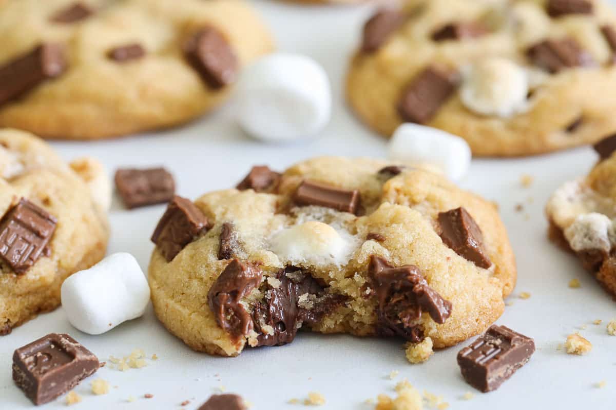 Horizontal image of a S'mores Cookie with a bite missing surrounded by more cookies and marshmallows and chocolate.