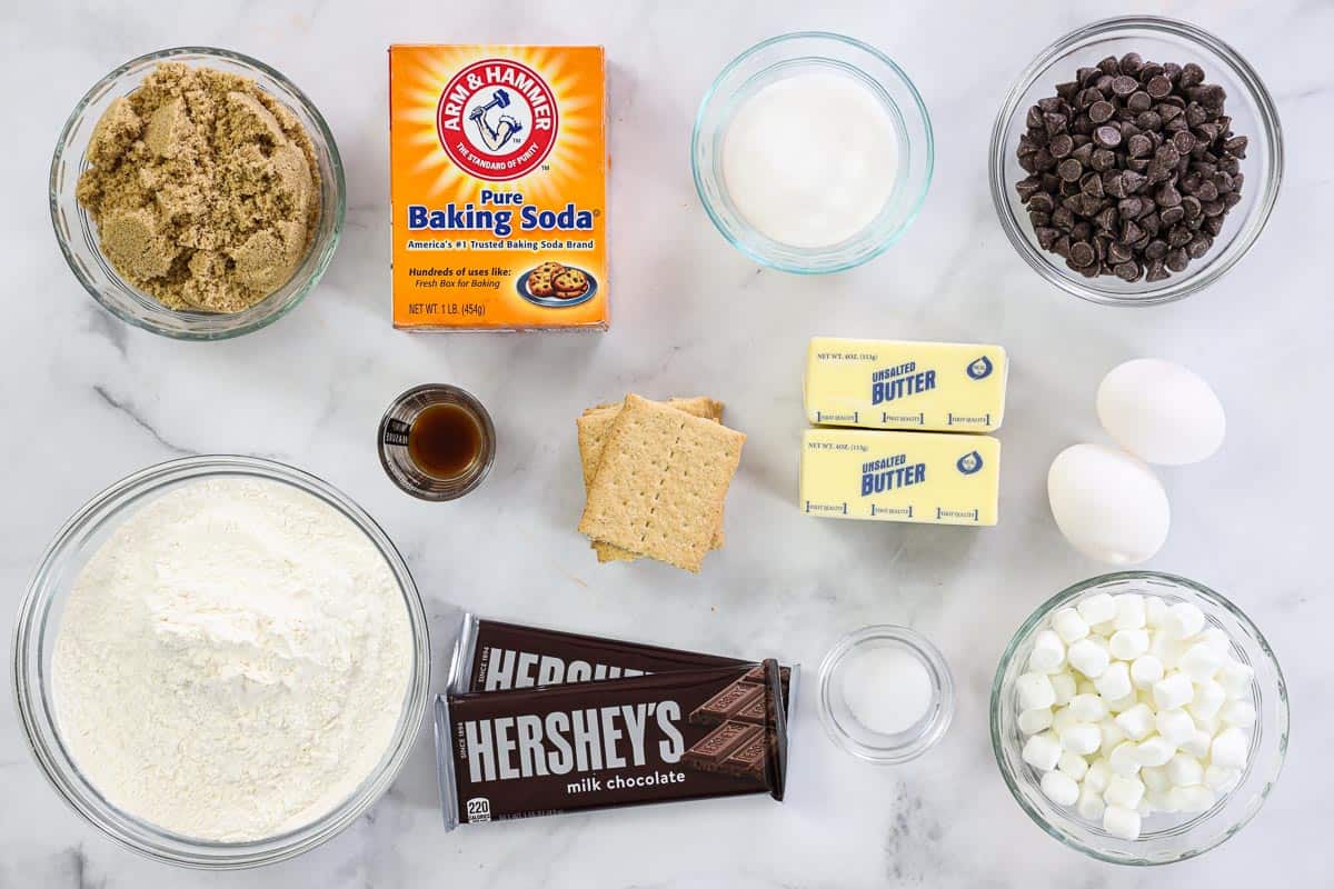 Ingredients for S'mores Cookies, including flour, sugar, brown sugar, butter, eggs, vanilla, salt, chocolate chips, mini marshmallows, Hershey's chocolate bars, and graham crackers.