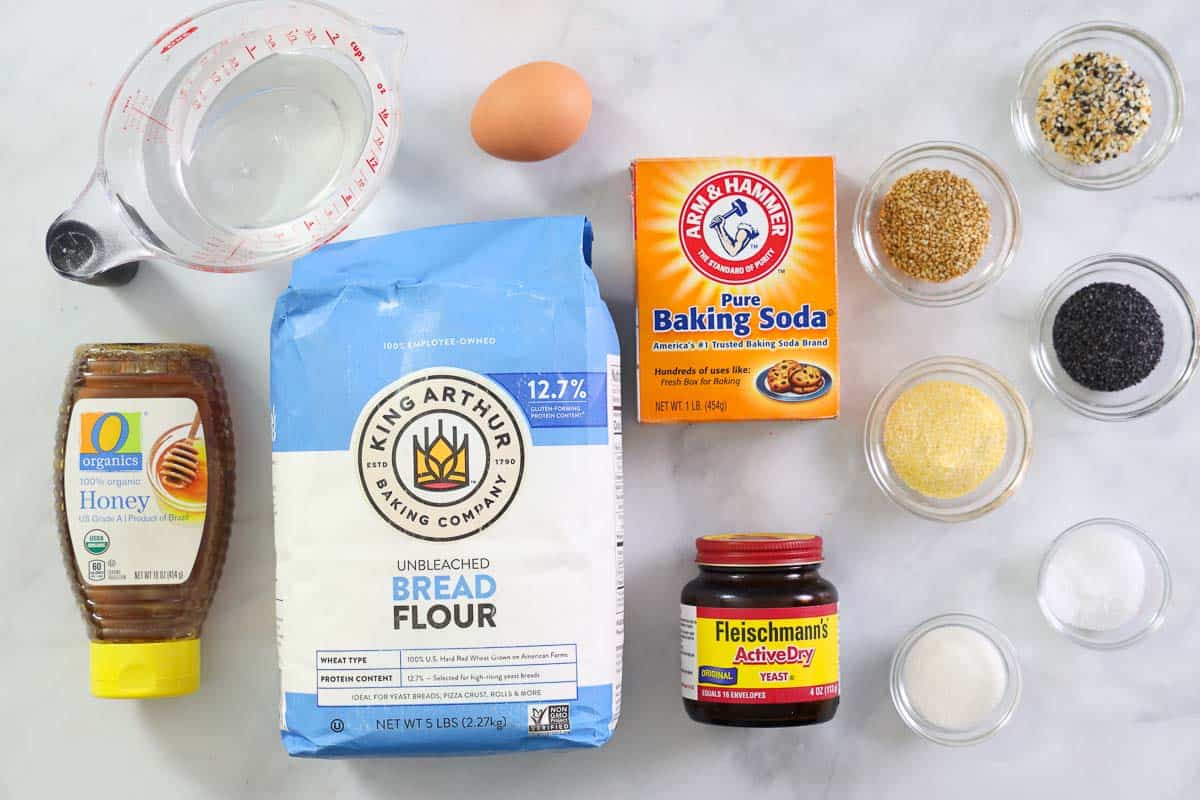 Ingredients for making New York Bagels, including bread flour, salt, water, yeast, sugar, honey, baking soda, corn meal, and toppings like sesame and poppy seeds and everything bagel seasoning mix.