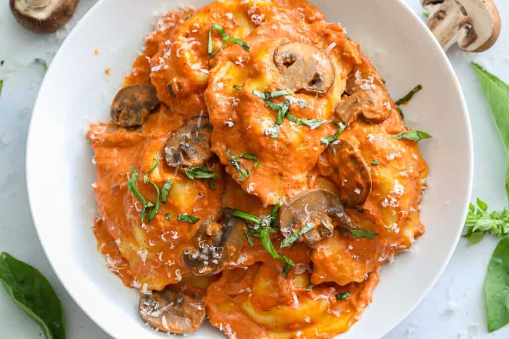 overhead image of Italian sausage filled ravioli in a white plate with fresh basil and mushrooms beside it.