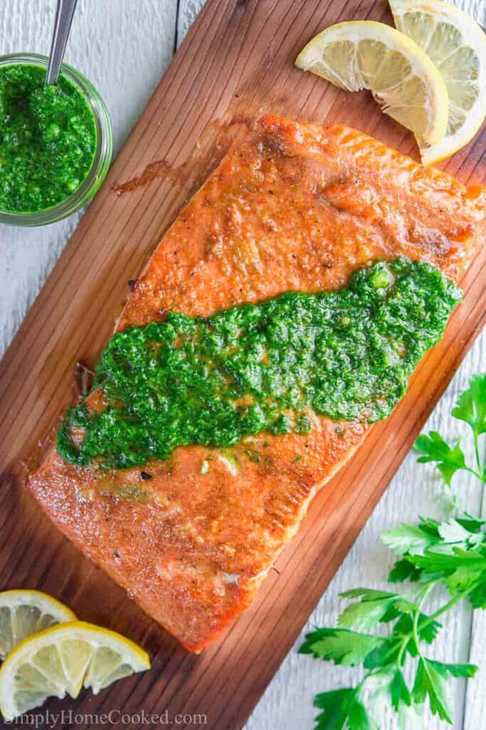 This easy grilled cedar plank salmon is full aromatic flavors from the garlic and spice rub. This recipe is served with homemade chimichuri sauce. #cedarplanksalmon #cedarplank #salmon #wildcaughtsalmon #salmonrecipe Vertical image of Cedar Plank Salmon with chimichurri sauce on top
