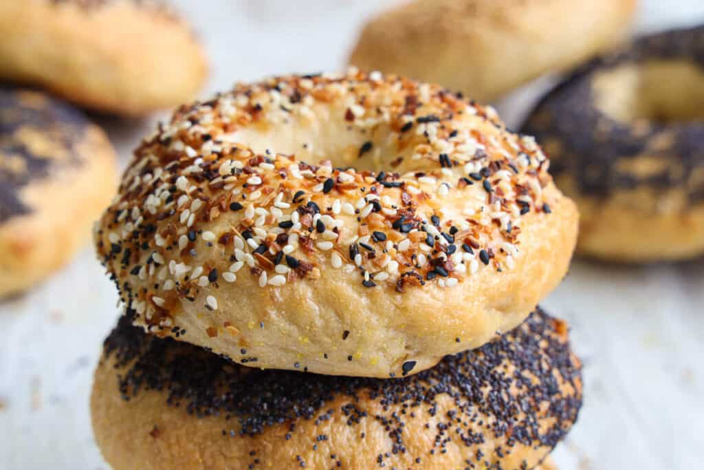 Everything Sourdough Bagel stacked on a poppy bagel, with more sourdough bagels in the background.